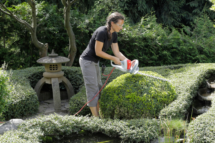 Eine Frau schneidet in einem gepflegten Garten einen Busch mit der Ultralight Heckenschere FHS 1545 in eine Kugelform.