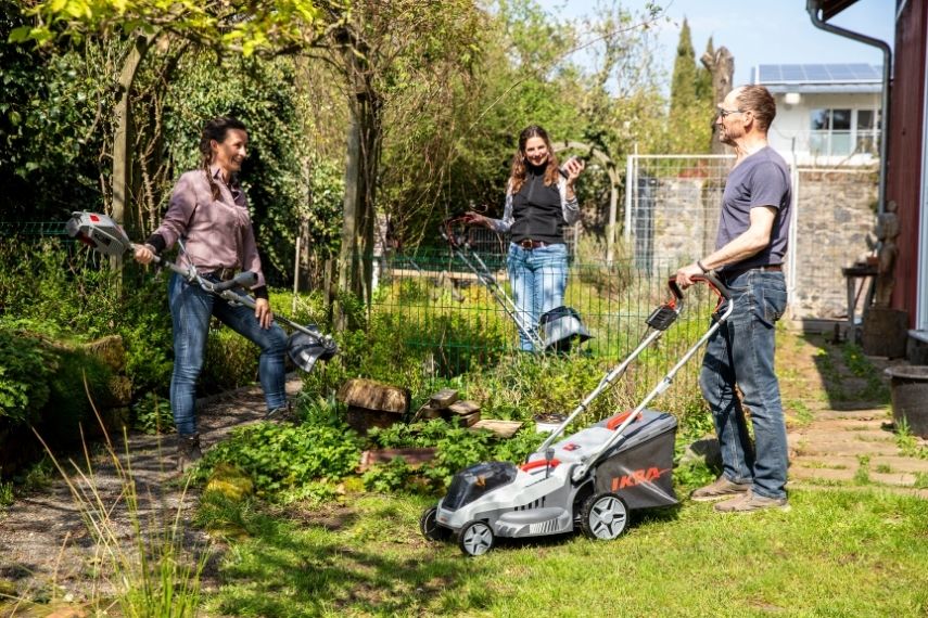 Drei Personen stehen an einem Gartenzaun und halten verschiedene IKRA x BOSCH Akku-Gartengeräte: die Bodenhacke IB-TC 36, den Rasenmäher IB-LM 33 und den Freischneider IB-BC 33.