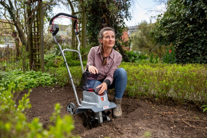 Eine Frau kniet im Beet neben der Akku Bodenhacke IB-TC 36 und setzt den Akku in das Gerät ein.