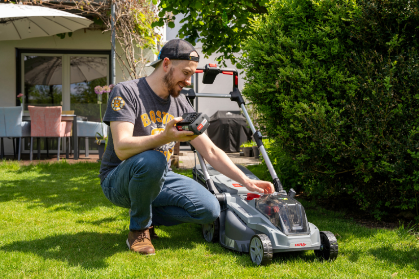 Ein Mann hält einen 36 Volt BOSCH Akku von IKRA in der Hand. Er kniet dabei neben IKRA Akku Rasenmähers IB-LM 43 auf einem grünen Rasen vor einem Einfamilienhaus.