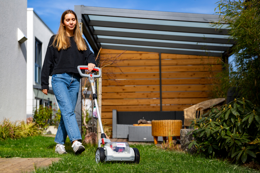 Einhandmäher ICHM 20 Eine junge Frau mäht im Garten mit dem kompakten Akku-Einhandmäher ICHM 20 von IKRA. Sie führt das Gerät mit einer Hand über eine Rasenfläche.